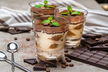 Classic tiramisu dessert in a glass cup and pieces of chocolate bar on concrete background