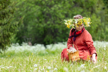 A girl in a national costume and a wreath on her head collects berries in a basket, on a green lawn in