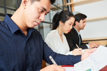 Obraz premium Asian young office worker sitting at the table examining financial graphs during work day at office with other colleagues sitting near by him and working