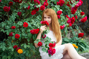 Portrait of a pretty redhead girl dressed in a white light dress on a background of blooming roses. Outdoor.