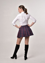 full length portrait of a brunette girl wearing a red leather jacket and plaid skirt, standing pose with back to the camera on a cream studio background.