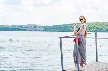 Girl in white T-shirt and skirt in sunglasses on small wooden pier on  shore of lake.