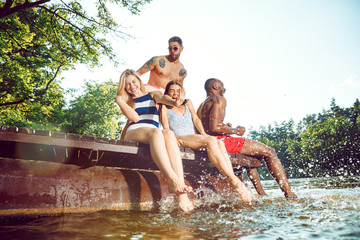 Fototapeta premium Group of happy friends having fun while sitting and laughting on the pier on river. Joyful women and men in swimsuit at riverside in sunny day. Summertime, friendship, resort, relations concept.