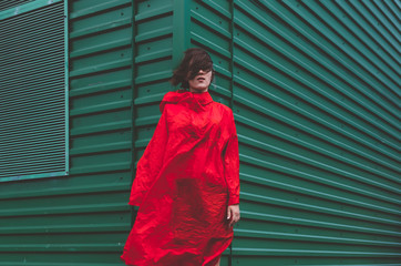 Cute positive short haired women in red rain coat, Standing over green industrial wall