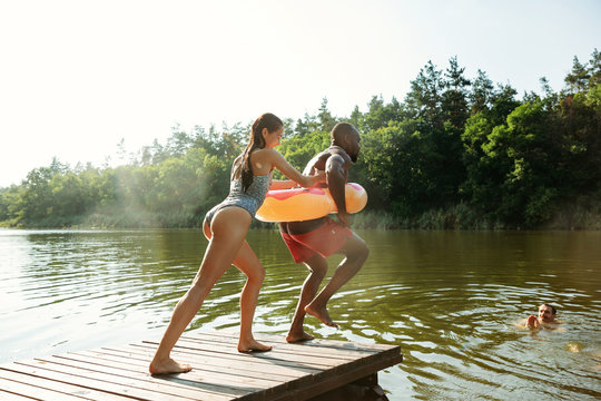 Happy friends having fun while laughting and jumping from the pier in river. Joyful male and female models in swimsuit at riverside in sunny day. Summertime, friendship, resort, weekend concept.