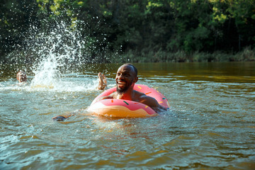 Happy man having fun while laughting and swimming in river. Joyful male models with rubber ring as a donut at riverside in sunny day. Summertime, friendship, resort, weekend concept.