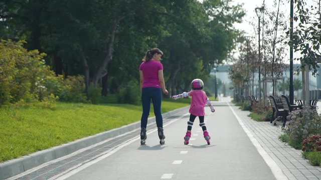 Little Girl with Mother Riding on Rollerblades Outdoors in Slow Motion. Young Woman Teaching Little Girl to Rollerblading in Sunny Day. Active Family Lifestyle Concept