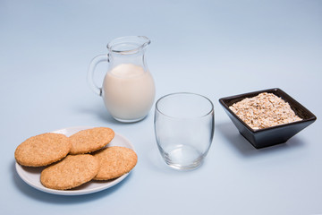 Leche de avena vegana sobre una mesa con un vaso de cristal, un bol con copos de avena y un plato de galletas.