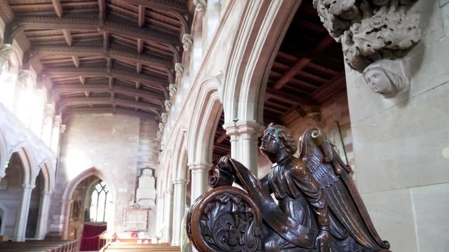Revelation Angel.  Camera tracks in to a wooden angle carved on a pew. Light fills the left of the frame.