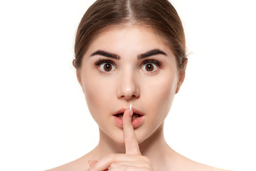 Close-up portrait of a beautiful young girl holding finger near her mouth expressing idea of secret isolated over white background.