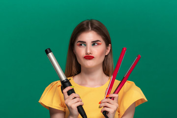 Portrait of beautiful young woman with bright color make-up choosing between curling and straightening irons for hair style.