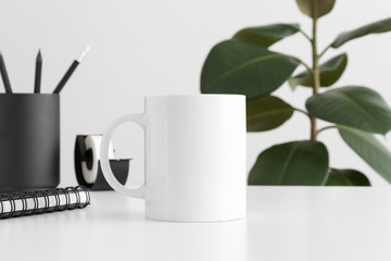White mug mockup with workspace accessories on a white table and a ficus plant.