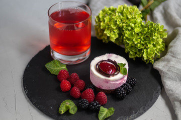 A glass with a compote of berries, next to a plate with blackberries, blueberries, raspberries. All this on a dark background, decorated with flowers.