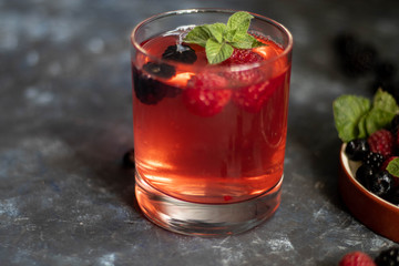 A glass with a compote of berries, next to a plate with blackberries, blueberries, raspberries. All this on a dark background, decorated with flowers.
