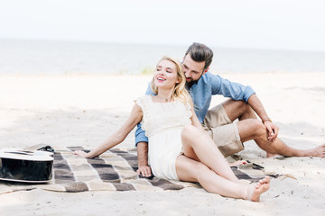 beautiful young barefoot couple sitting on blanket near acoustic guitar at beach near sea