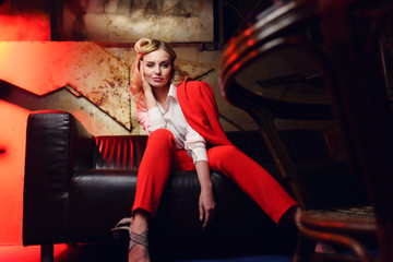 Fototapeta na wymiar Full-lenght photo of young blonde woman in red jacket looking at camera sitting on leather sofa