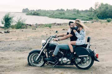 young couple of bikers hugging on black motorcycle at sandy beach near river