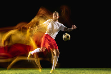 In fire. Young caucasian male football or soccer player in sportwear and boots kicking ball for the goal in mixed light on dark background. Concept of healthy lifestyle, professional sport, hobby.