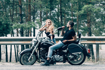 Plakat young couple of bikers sitting on black motorcycle and man drinking alcohol from bottle on road near green forest