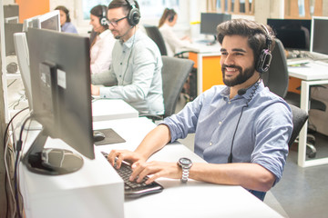 Friendly handsome young man with hands-free headset working on computer in call centre