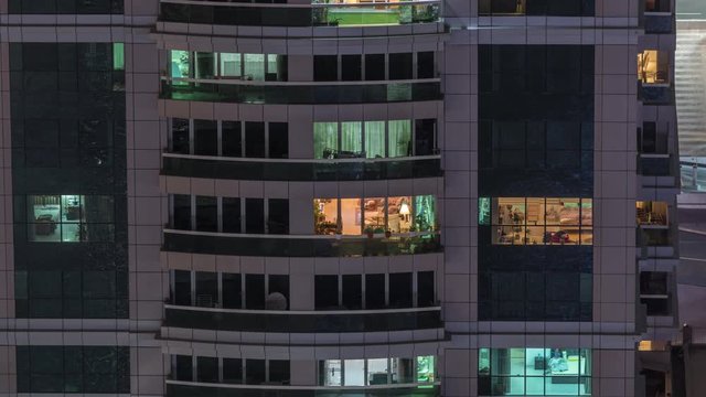 Night view of exterior apartment building timelapse. High rise skyscraper with blinking lights in windows with people moving inside. Pan down