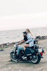 young loving couple of bikers sitting on black motorcycle at beach near river