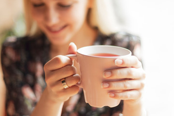 Close up photo of pretty woman holding mug of delicious hot tea, drinking, smiling, in cozy morning...