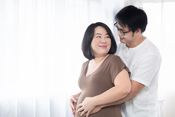 Lovely Asian adult pregnant couple happy hugging together with hand of husband touching belly of his wife