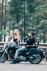 Obraz na płótnie Canvas side view of young couple of bikers sitting on black motorcycle while man drinking alcohol from bottle on road near green forest