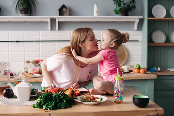 Obraz na płótnie Canvas Side view of cute mother and daughter hugging and kissing each other in morning. Family sitting at kitchen, cooking breakfast and cutting vegetables at home. Concept of lunch and food.