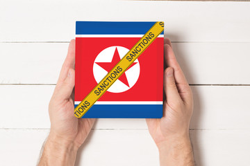 Yellow tape with the inscription Sanctions on the box with the flag of Noth Korea in male hands....