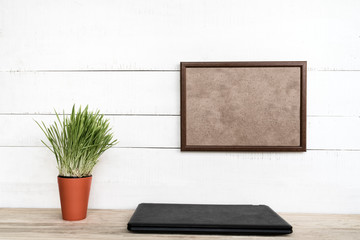 Empty frame on white wall. Closed notebook and green houseplant. Home workplace. Place for text