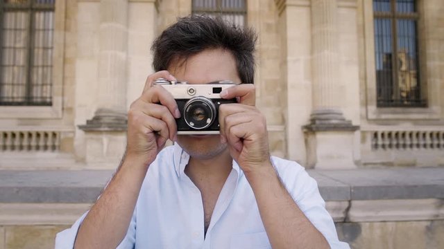 PARIS, FRANCE, APRIL 2019. Gimbal slow motion portrait shot of smiling young man in white shirt and eyeglasses with a film camera making a photo straight at camera on background of Louvre museum