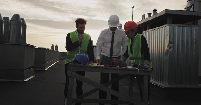 Three construction workers surround a desk looking at blue prints of the construction , they all point at the board looking interested, one ravishing woman with afro like hair and a handsome man with