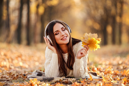 Beautiful woman with headphones and maple leafs lying on the ground in autumn park