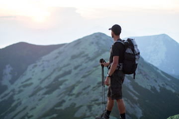 caucasian man hiker with backpack and trekking sticks is on the peak of green stones mountain enjoy a panoramic view of mountains in the sunset light