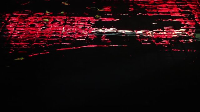 Rainy night. Beautiful red street lighting reflected in rainwater puddle on the road paved wth setts. Rain drops creating beautiful ripples, texture and rhythmical abstract patterns.