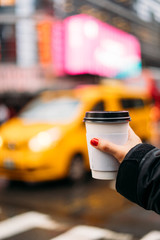 Hand of a girl holding a white coffee glass with a yellow taxi out of focus in the background