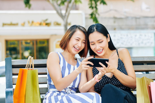 Two Asian young women resting on bench after shopping and watching funny photos on mobile phone together in the city