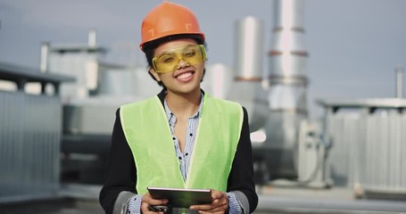 Good looking African ethnic young woman architect looking straight to the camera and smiling while holding a ration and tablet , and have a safety equipment - Powered by Adobe