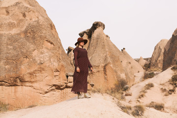 Travelling woman wearing brown hat and long dress walking in sand valley and rocks. Travel and wanderlust concept. Cappadocia, Turkey.
