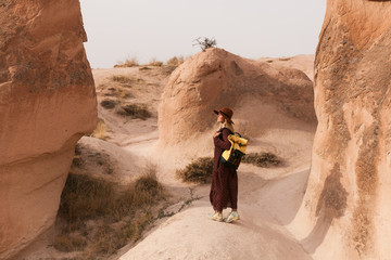 Travelling woman wearing brown hat and long dress walking in sand valley and rocks. Travel and wanderlust concept. Cappadocia, Turkey.