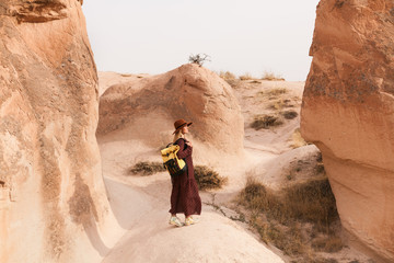 Travelling woman wearing brown hat and long dress walking in sand valley and rocks. Travel and wanderlust concept. Cappadocia, Turkey.