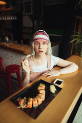Vertical photo of attractive girl in pink clothes and cap, sitting at table in restaurant with chopsticks, eating sushi, and looking into camera with astonished face. Japanese cuisine concept.