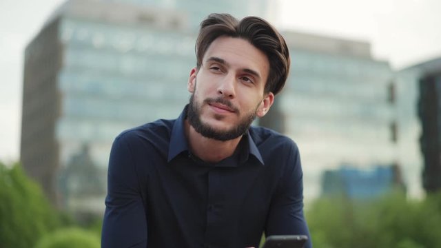 Zoom in of bearded young businessman sitting on bench in park, using cell phone and looking away dreamily
