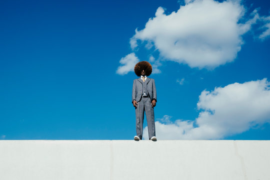Portrait confident, cool, well-dressed young man with afro standing on wall against sky