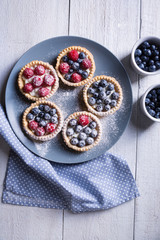 Cakes with raspberries and blueberries on a gray large plate sprinkled with icing sugar on a white wooden background Top view