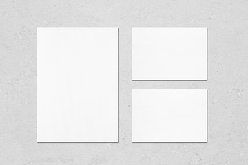 One empty white vertical a4 sized poster and two a5 sized horizontal rectangle card mockups with soft shadows on neutral light grey concrete wall background. Flat lay, top view