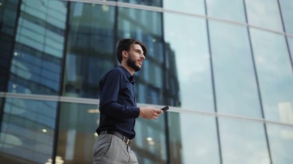 Panning slow motion shot of confident and handsome young businessman walking down modern city street and looking away thoughtfully. Man holding cell phone and typing sms - Powered by Adobe
