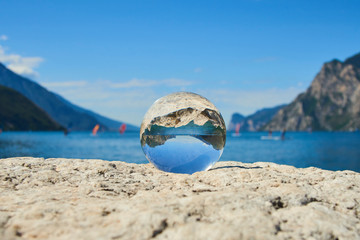 Fototapeta na wymiar Lake Garda (Lago di Garda or Lago Benaco) seen through a glass crystal ball placed on rock. Selective focus